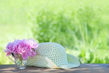 Pink peonies flowers in glass cup and braided hat in garden, sunny natural background. Summer time...