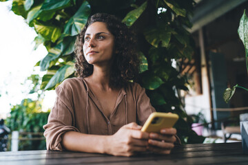Contemplative female blogger with modern cellular gadget resting at cafe terrace thinking about web...