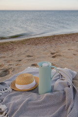Straw hat and thermos on the blanket against a sand beach and blue sea. Romance of the dawn. Morning on vacation. Picnic on the beach. Summer headgear. Women’s hat