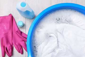 Basin with white garment near detergent and gloves on floor, flat lay. Hand washing laundry
