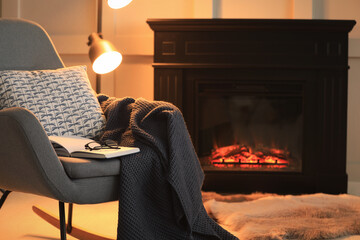 Open book, glasses and plaid on rocking chair near fireplace at home, space for text. Cozy...