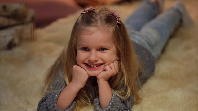 Charming little girl lying on soft carpet at home smiling looking away. High angle view portrait of pretty Caucasian kid enjoying domestic leisure indoors. Slow motion