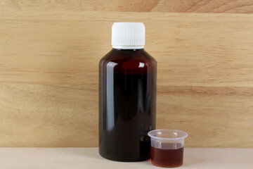 Bottle and measuring cup with cough syrup on white table against wooden background