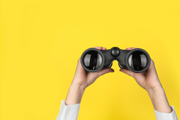 Woman holding modern binoculars on yellow background, closeup. Space for text