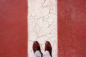 POV: a man looking at his feet while standing on a striped red and white asphalt surface. Overhead shot.
