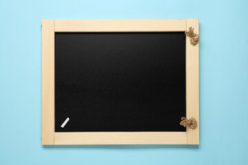 Clean small black board with piece of white chalk on light blue background, top view