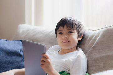 Cinematic portrait young boy playing game on tablet sitting on sofa with light shining from window, Kid playing games online on internet at home, Child talking video call with friends at home