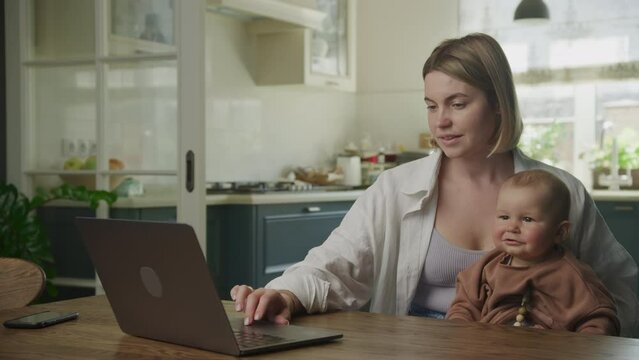 Young adult caucasian mother holding her son and working on laptop in joy