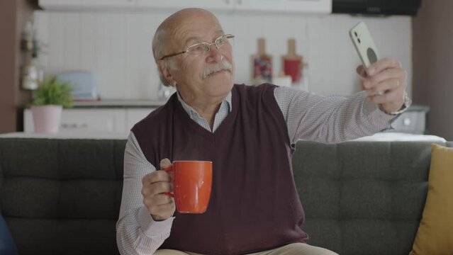 Happy 70s male user making video call online while drinking coffee, looking at screen sitting on sofa at home, taking selfie, old grandfather learning to use modern technology.