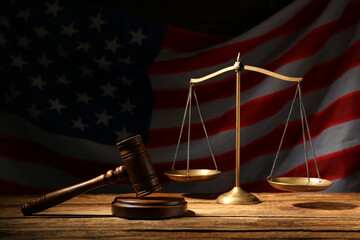 Scales of justice and judge's gavel on wooden table against American flag in darkness