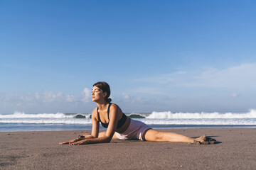 Flexible fit girl practice twine stretching during morning workout at coastline seashore beach, good looking female in sportswear enjoying healthy lifestyle for body care warming up muscles in asana