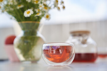 A glass teapot and a cup of chamomile tea on the background of a vase with daisies. Morning breakfast in the fresh air. High quality photo