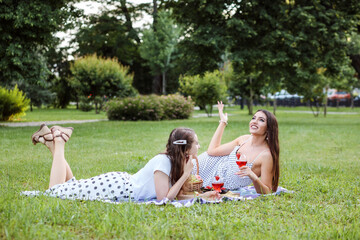 Two women having picnic together, laying on the plaid on the park lawn and drinking cocktails in summer