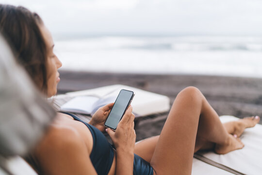 Millennial female tourist using cellular technology for chatting and messaging in social media networks during recreation time at beach coastline, young woman writing content mobile publication