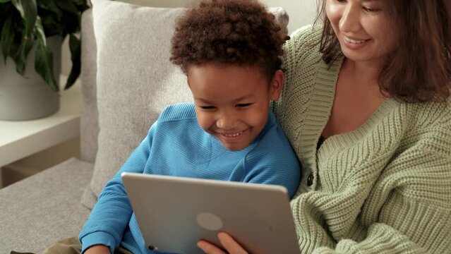 Close up view of asian mom and her cute african son sitting on sofa, watching cartoons together at tablet, making wide beaming smile and laughing, having a good time. 4k footage