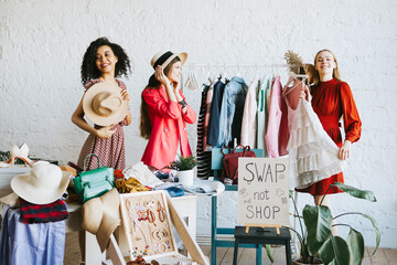 three young woman female caucasian and african students at swap party try on clothes, bags, shoes...