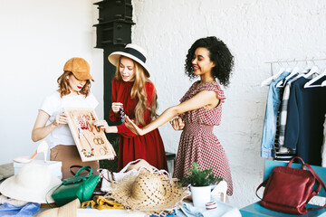 three young woman female caucasian and african students at swap party try on clothes, bags, shoes...