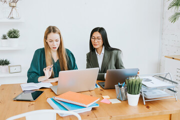 two young business women professionals in formal wear clothes work in modern office using laptop, tablet, brainstorm and search for solutions together, confident independent Asian girl solves problems