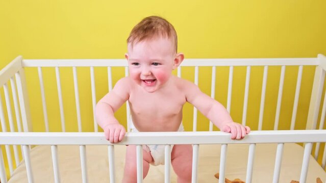 Happy infant baby boy is standing in a crib, yellow studio background. Smiling caucasian toddler kid in diaper