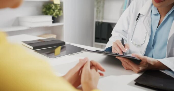 Close up of Young Asia female doctor in white medical uniform using clipboard is delivering great news talk discuss results or symptoms with girl patient sitting at desk in health clinic office.