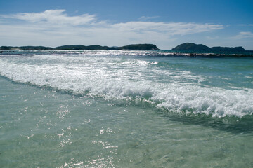 Fototapeta na wymiar Lindas ondas em praia de areias brancas, céu azul com nuvens e montanhas, localizada na região de Cabo Frio, Rio de Janeiro, Brasil.