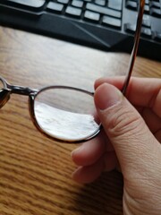 A man's hand holding glasses on a wooden desk with a computer keyboard.