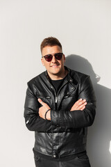 Obraz premium Handsome hipster cool man with smile in black fashion leather jacket and sunglasses stands near a gray wall