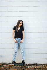 A teenage girl with long hair wearing black, denim and combat boots leaning against an old cinder...
