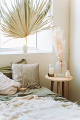 Interior of sunny modern bedroom with bed covered with grey bed linen, beige plaid. Big glass vase with dry pampas, decorative candle on bedside table. Huge palm leaf in white vase on windowsill. 
