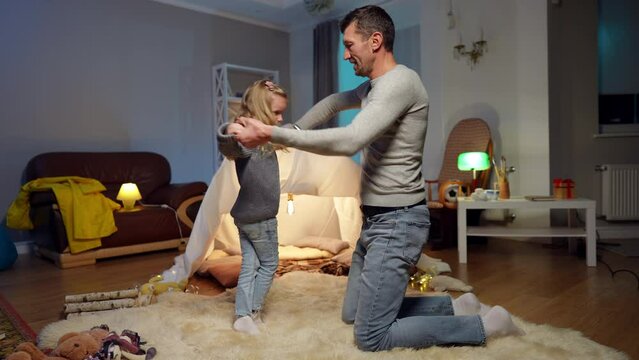 Wide shot happy daughter dancing with father standing on knees at tent in living room. Portrait of carefree smiling cute Caucasian girl enjoying dance with positive man at home indoors