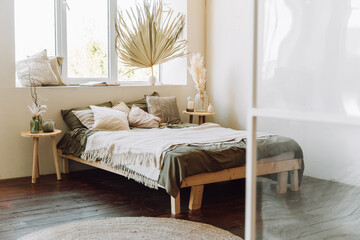 Interior of sunny bright modern bedroom. Wooden bed covered with grey bed linen, beige cotton plaid with fringe on brown wooden floor. Different glass vases with dry plants on wooden bedside tables.
