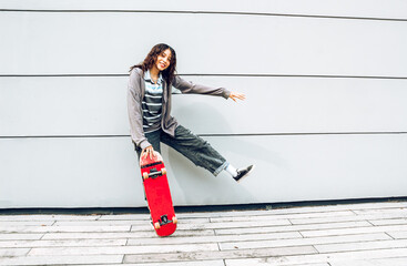 mujer morena con crespos divirtiéndose en la ciudad sosteniendo patineta roja y levantando un pie feliz en fondo de color de pared de edificio 
