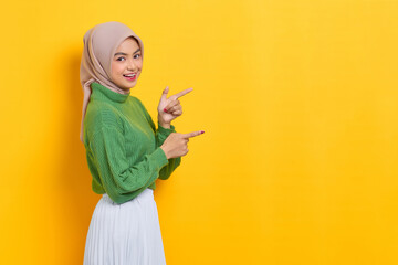 Cheerful beautiful Asian woman in green sweater pointing fingers aside at copy space isolated over yellow background