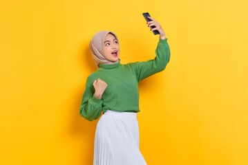 Excited beautiful Asian woman in green sweater holding mobile phone and celebrating great news message isolated over yellow background