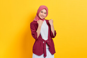 Excited beautiful Asian woman in casual shirt celebrating big luck and success with raised fists isolated over yellow background