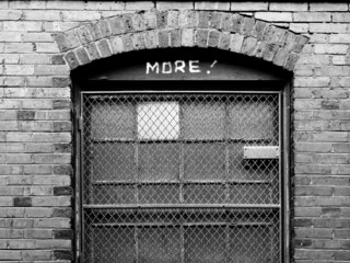 black and white of an old brick factory building with iron bars on window and word MORE spray...