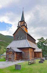 Fototapeta na wymiar Kaupanger Stave Church in western Norway. Old wooden walls and bell tower, cloudy grey sky, green grass and trees, stone tombs of the old cemetery, mountain slopes on the background