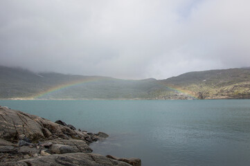 Rainbow in the cloud above the  Styggevatnet glacier lake in Norway. Rocky shore and foggy mountains around
