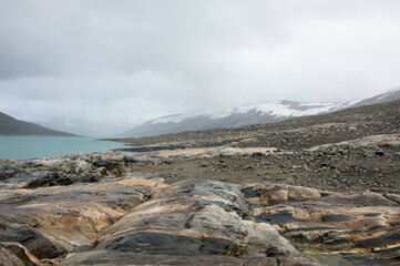 Shore of the Styggevatnet glacier lake in Norway. Turquoise water, grey and brown stones, snor on the mountain slopes,  cloudy sky