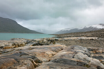 Shore of the Styggevatnet glacier lake in Norway. Turquoise water, grey and brown stones, snor on the mountain slopes,  cloudy sky
