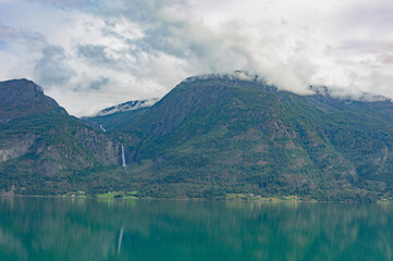 View of the Lustrafjord in Norway.  turquoise sea water, low white clouds above it, cloudy sky, green forest on the mountain slopes, white waterfall 