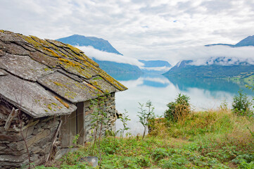 Rural landscape in Norway. Sea shore, old boathouse, covered with mossy stone tiles, old road, white cloud above the water in the Lustrafjord