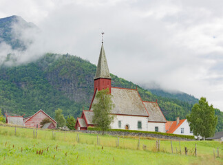 Fototapeta na wymiar Medieval 12 century gothic stone Dale church in Luster village on Lustrafjord in Norway. White plastered stone walls, green grass and mountain slopes, cloudy sky