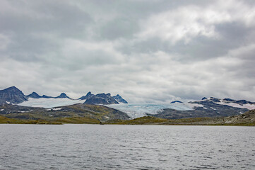Norwegian mountain landscape with snow covered melting  glacier, lake and grey sky with clouds.  - Powered by Adobe