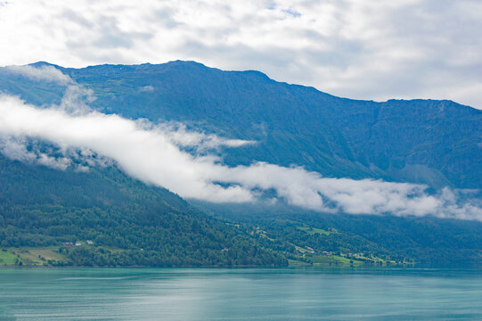 View of the Lustrafjord in Norway. Blue sea water, low white clouds above it, cloudy sky, green forest on the mountain slopes