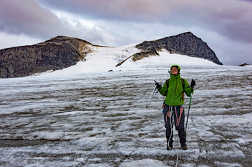 young woman alpinist standing near the crack in the glacier on the way to the Galdhopiggen mountain summit in Norway. White and grey snow and ice, blue sky with clouds and dark mount on the background
