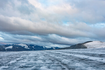 Evening on the glacier on the trail from Galdhopiggen summit in Norway. White and grey ice, dark mountains, sunset sky with clouds