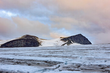 Galdhopiggen mountain - the highest summit of Norway. Blue sky with clouds, dark grey rocky slopes, white snow and ie of the glacier