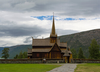 Fototapeta na wymiar Lom viking medieval stave church in Lom Norway. Old wooden walls and tower, green lawn and grey stone fence, mountain slope and blue sky with clouds on the background