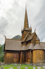 Fototapeta na wymiar Lom viking medieval stave church in Lom Norway. Old wooden walls and tower, green lawn and grey graveyard tombstones, mountain and cloudy sky on the background